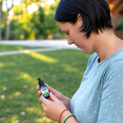 Femme dans un parc lisant l’étiquette d’un flacon d’huile de CBD, t-shirt bleu clair à pois, bracelets rouges et verts